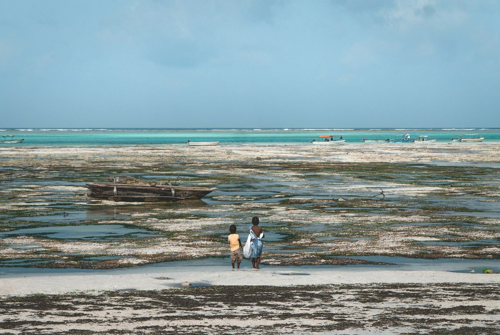 Jambiani beach at low tide — local children walking, traditional wooden boat, seaweed flats, turquoise horizon