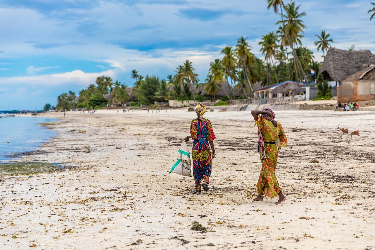 Women walking on Jambiani beach — colorful traditional dresses, white sand, palm trees, coastal village life